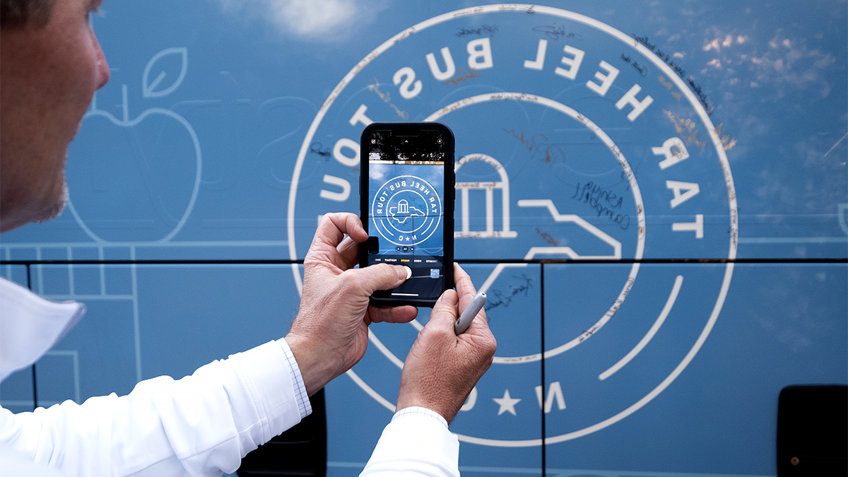 A man taking a photo on an iPhone of the side of a bus that features the logo of the Tar Heel Bus Tour on its side.